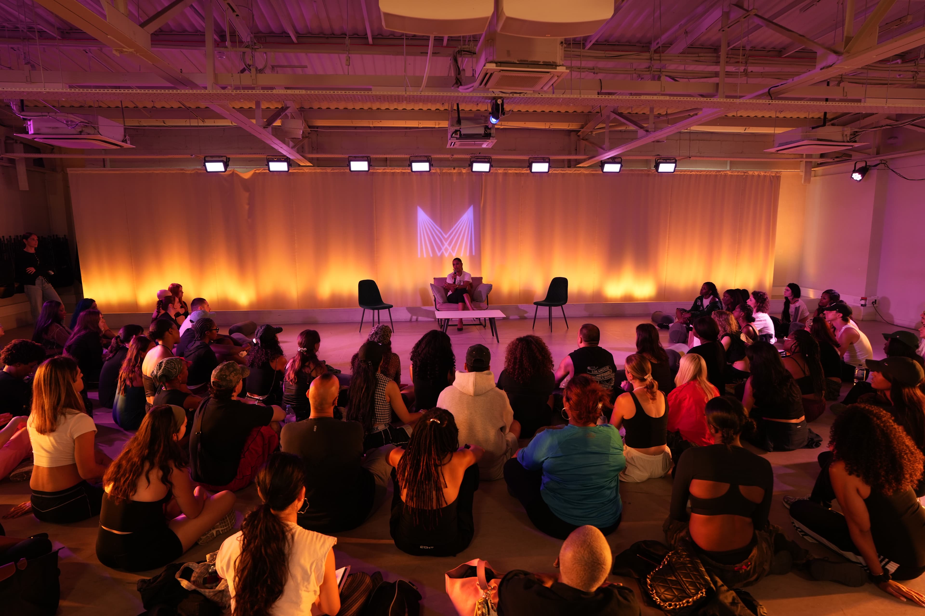 Victoria Monet giving a Q&A at The Manor LDN dance studio. The lighting is red and amber dimly lit with rustic style seating