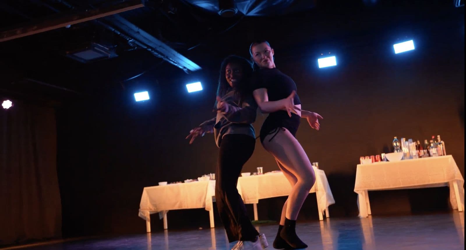 Two dancers mid-routine under blue studio lights, with party tables set behind them