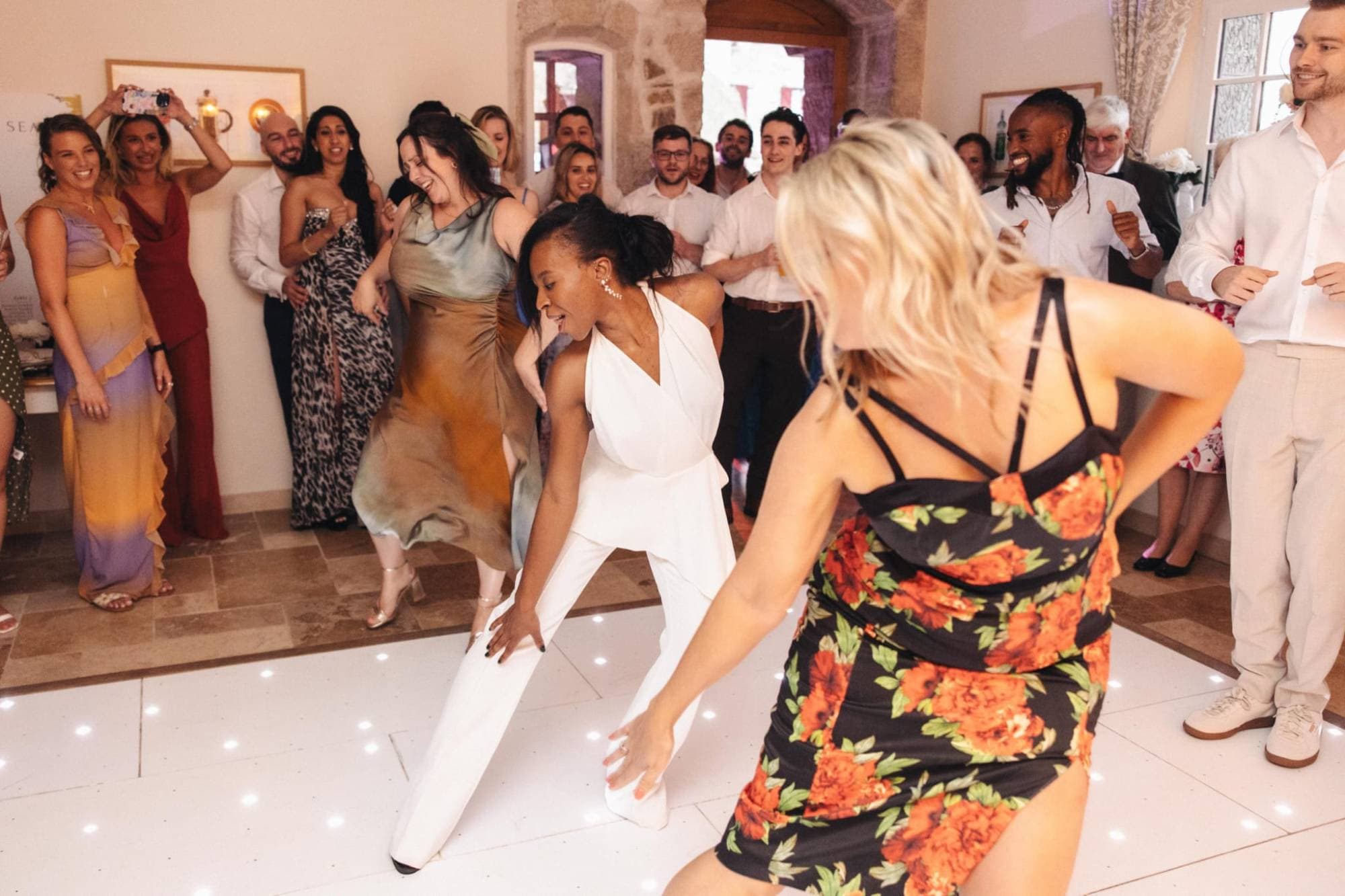 A bride in a white jumpsuit dancing a routine with her friends while wedding guests cheer and film