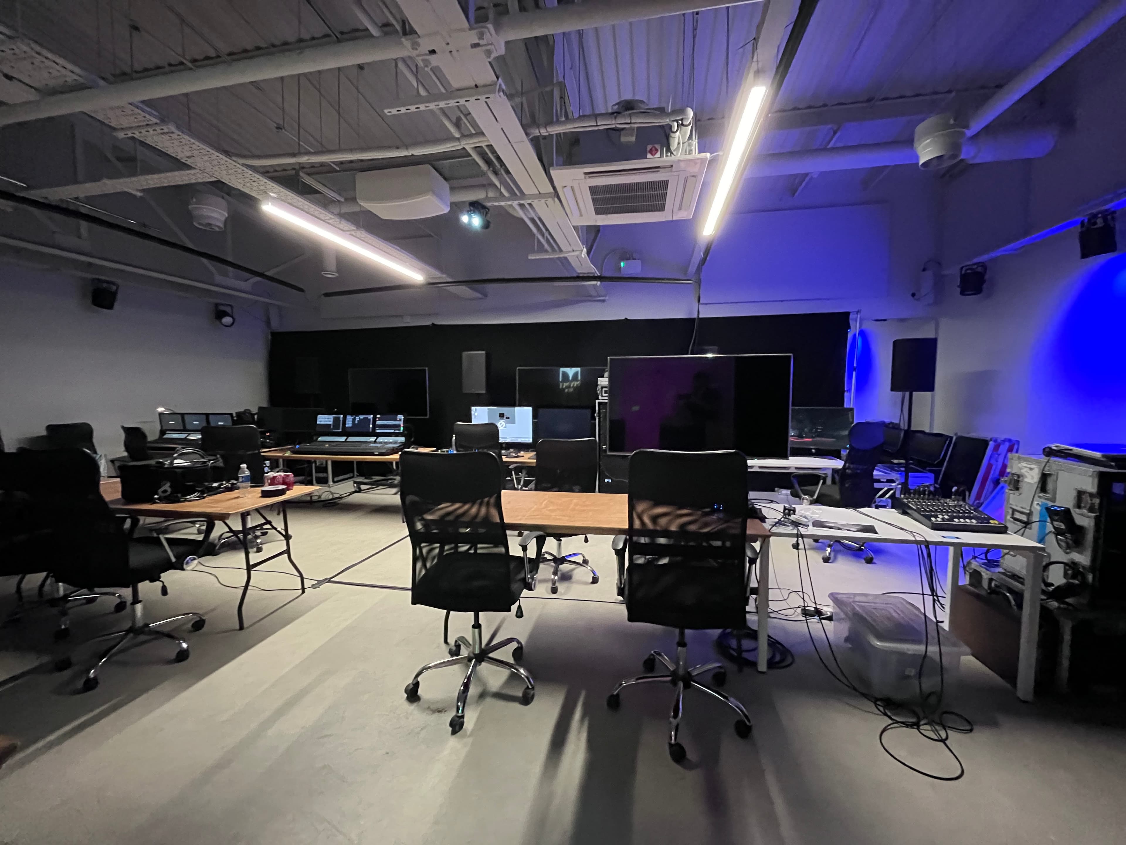 roduction studio set-up with multiple workstations, monitors, lighting and AV equipment, desks and office chairs arranged across the space, with blue production lighting illuminating the right side of the room.