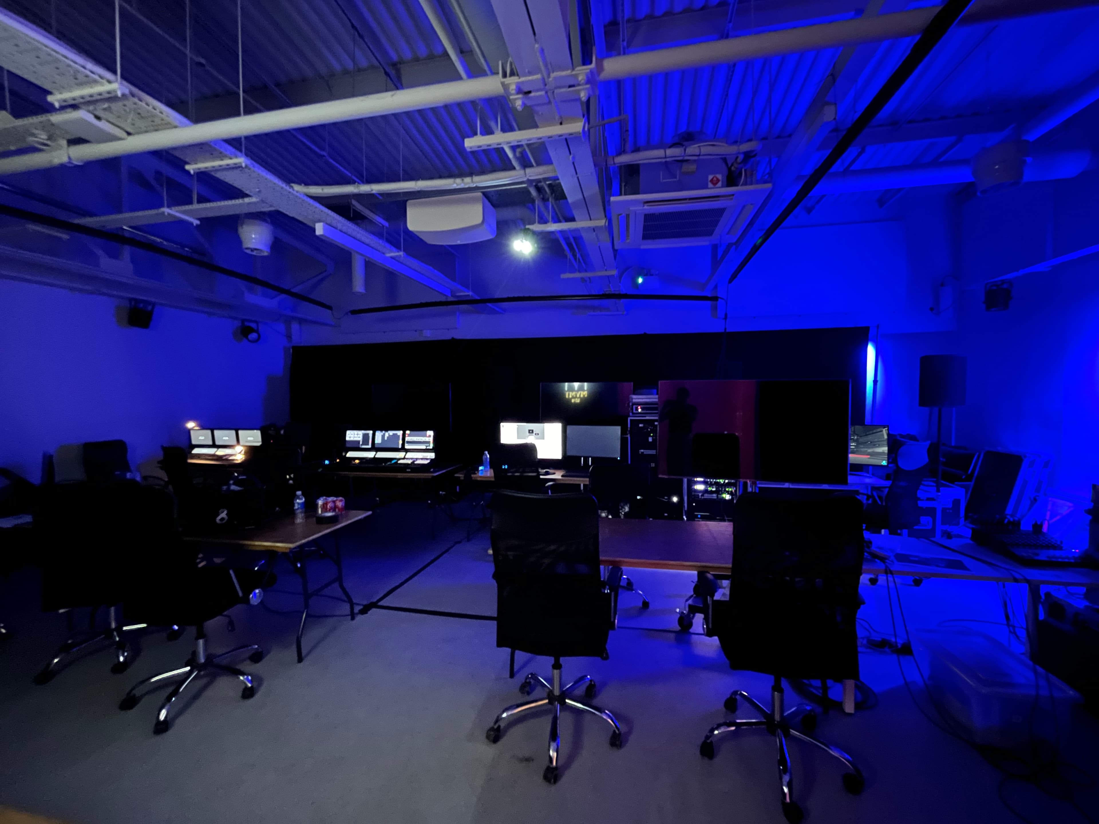 Production studio set-up with multiple workstations, monitors, lighting and AV equipment, desks and office chairs arranged across the space, with blue production lighting illuminating the right side of the room.